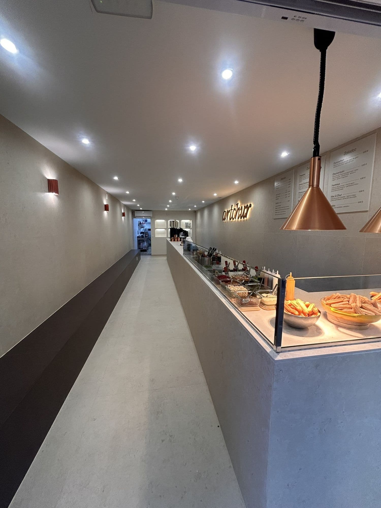 Interior de la churrería Artchur: barra corrida de piedra clara, lámparas cobrizas y rótulo retroiluminado al fondo