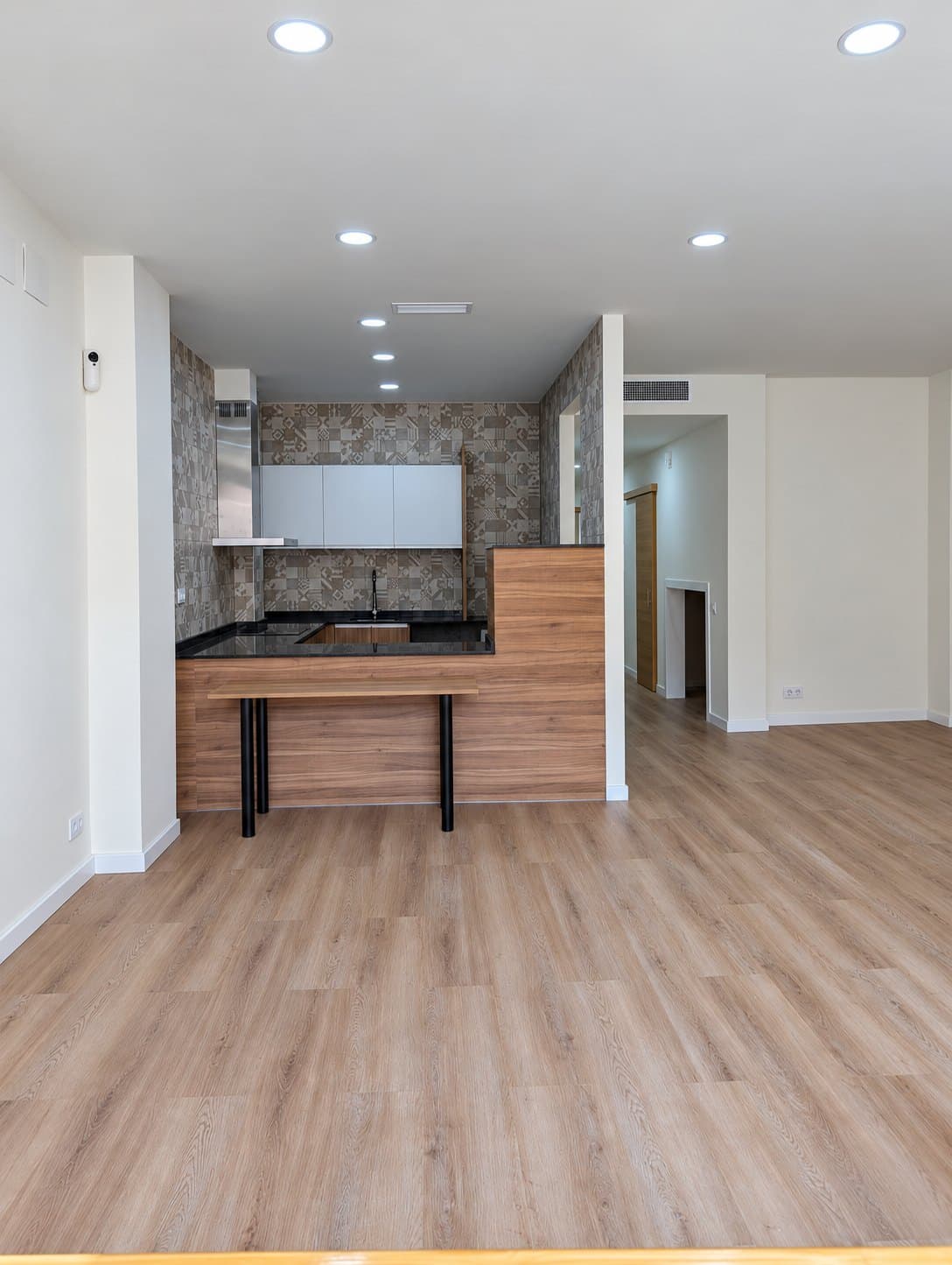 Salón de la vivienda accesible con la cocina abierta al fondo, barra baja y suelo de madera continuo