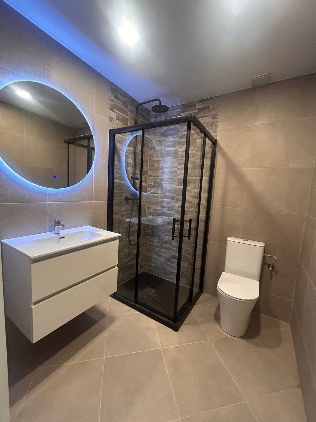 Baño con espejo redondo retroiluminado, mueble blanco y ducha con mampara negra