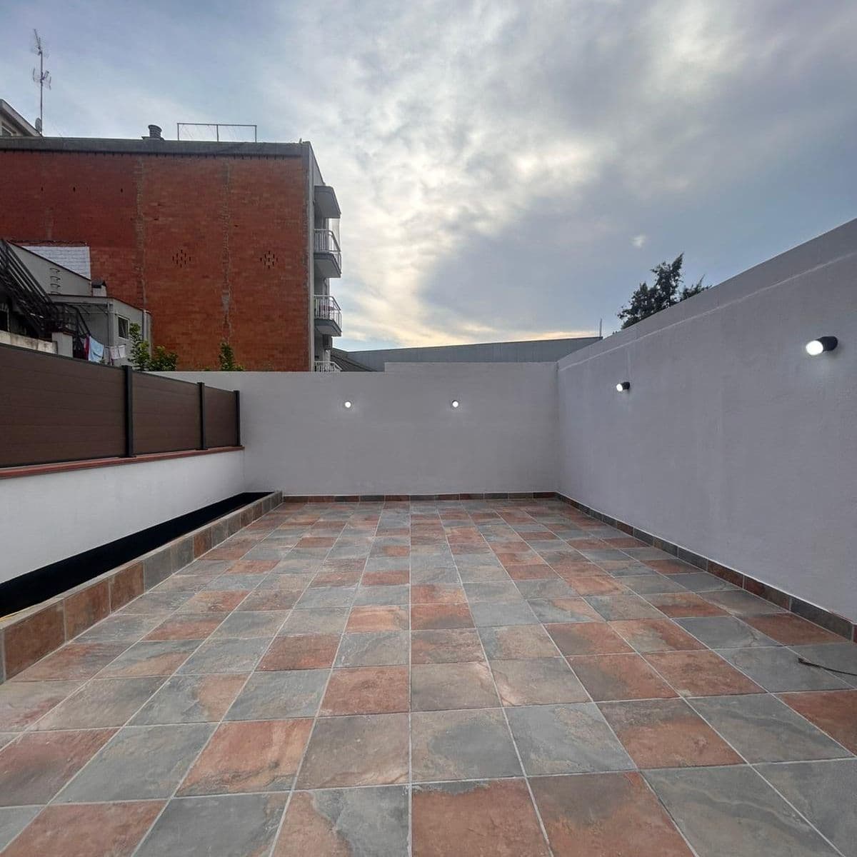 La misma terraza reformada: gres en tonos tierra, frente de lamas y apliques
